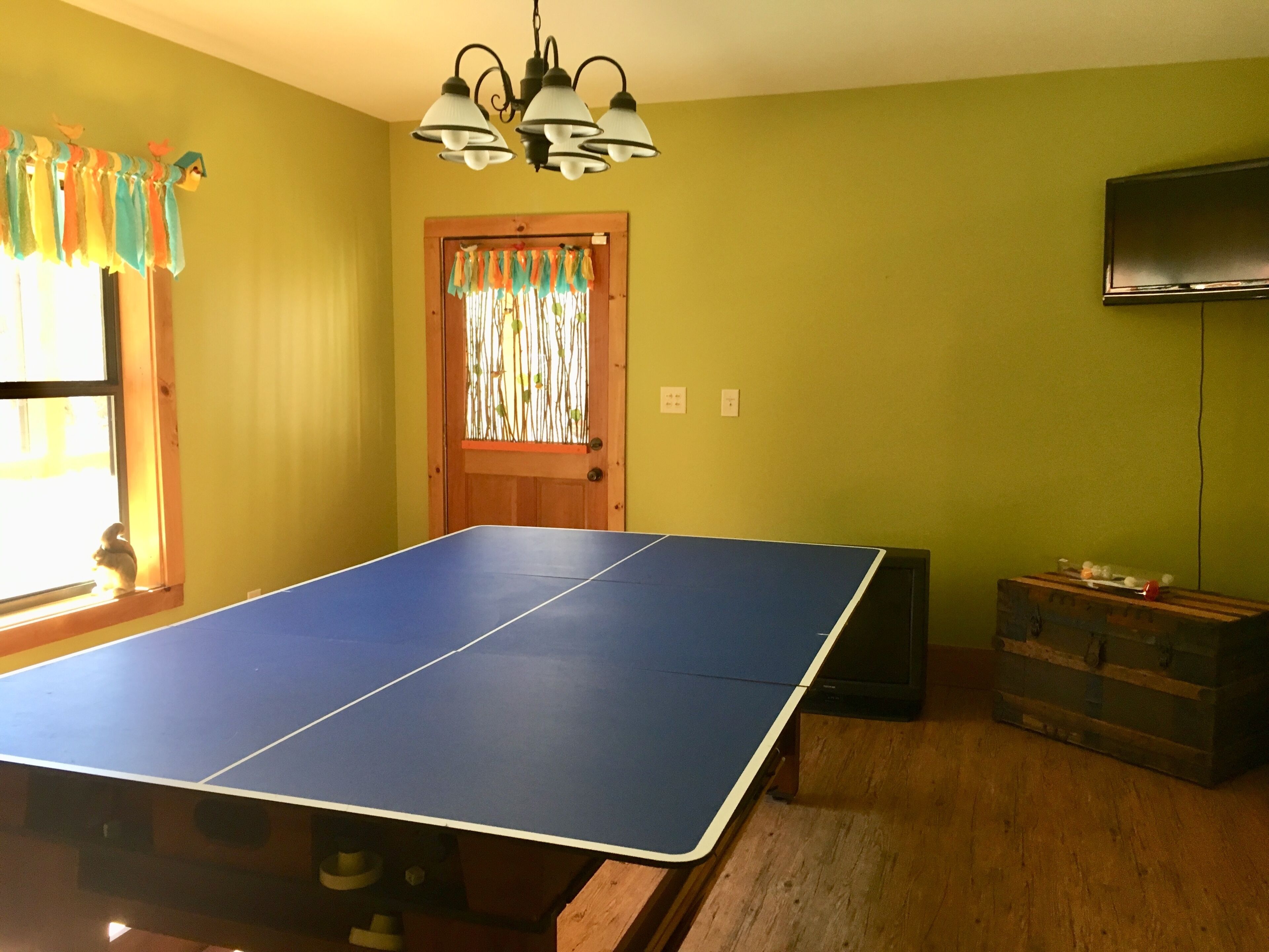 A room with a ping pong table, wooden flooring, a television on the wall, and a door with a colorful curtain.