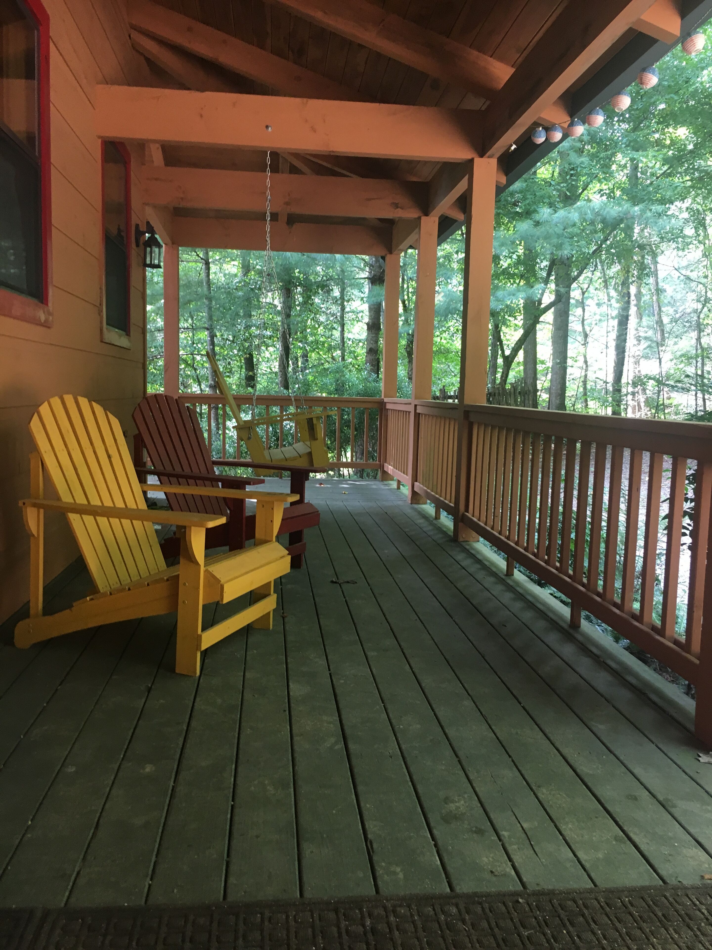 A wooden deck with Adirondack chairs and a table, surrounded by a wooden railing and trees.