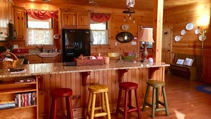 Interior de una cabaña de madera con cocina, que incluye un refrigerador, taburetes de bar de madera y una zona de comedor con banco.