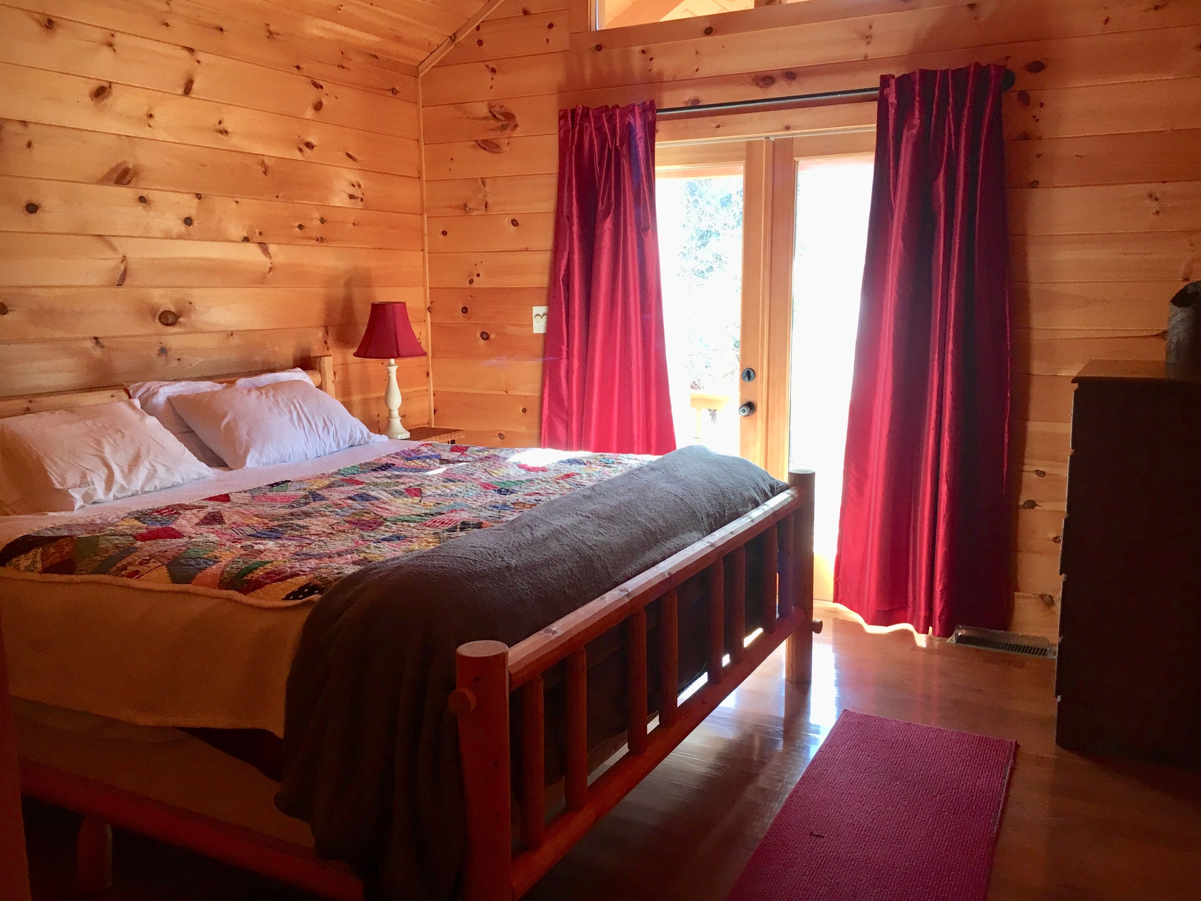 A wooden cabin bedroom with a bed, pillows, a quilt, a nightstand with a lamp, and a door with curtains.
