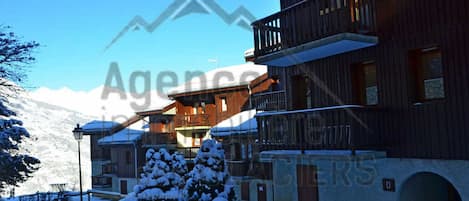 A snow-covered building with a balcony, a snow-covered tree, and a mountain in the background.