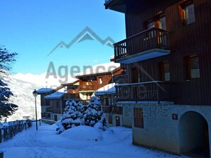 Un edificio cubierto de nieve con un balcón, un árbol nevado y una montaña al fondo.