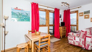 A cozy room with a wooden table and chairs, a sofa, and a view of mountains through large windows with red curtains. - Divisible studio near slopes, 4 pers., parking, nice view (Bellentre)