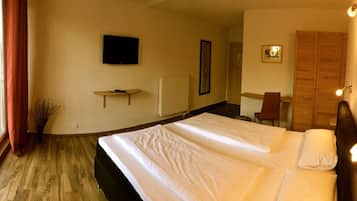 A hotel room with a large bed, a television on the wall, a wooden wardrobe, and a small table with a chair.