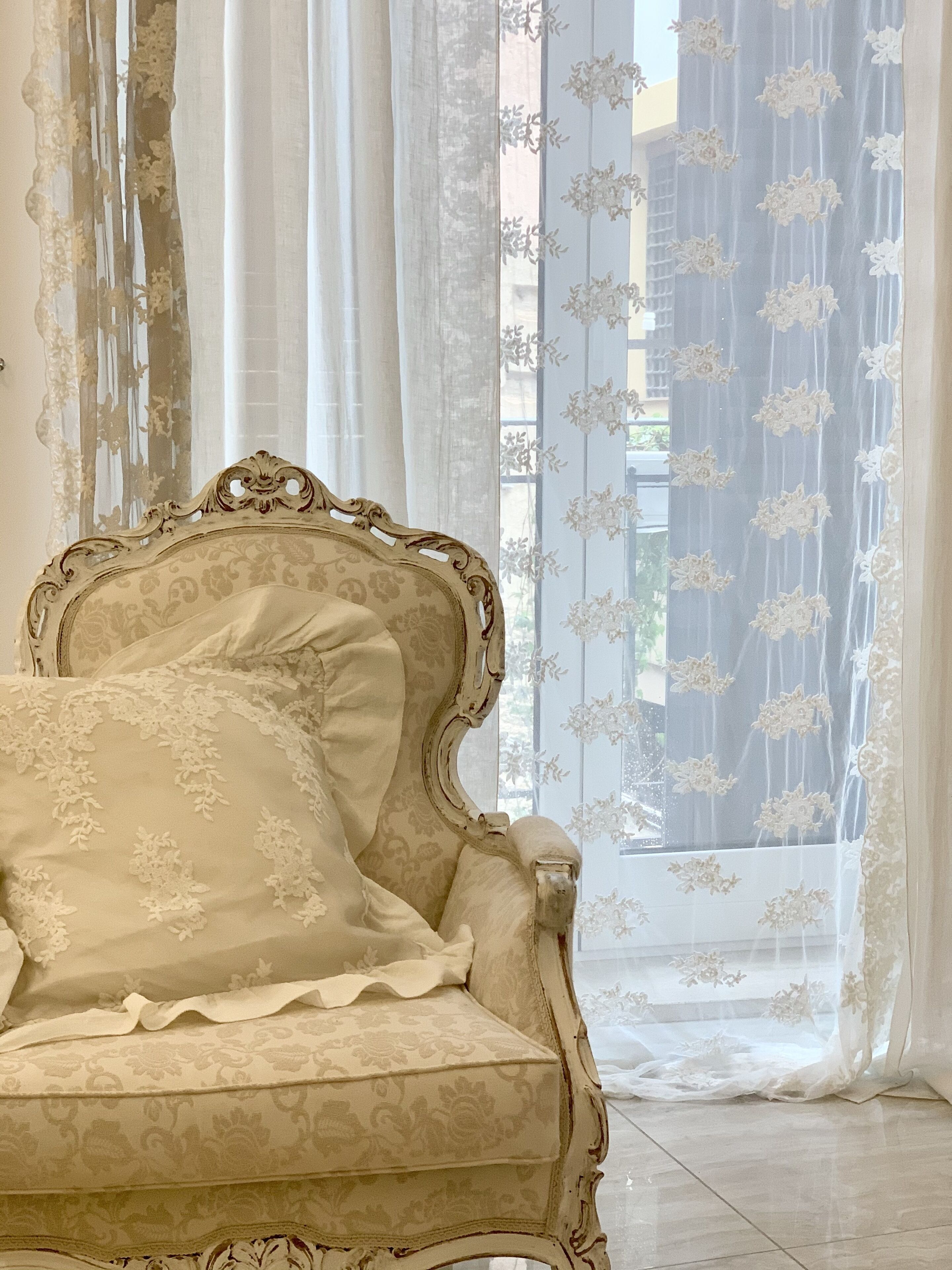 A vintage-style armchair with ornate detailing, positioned in front of a large window with sheer curtains and floral patterns.