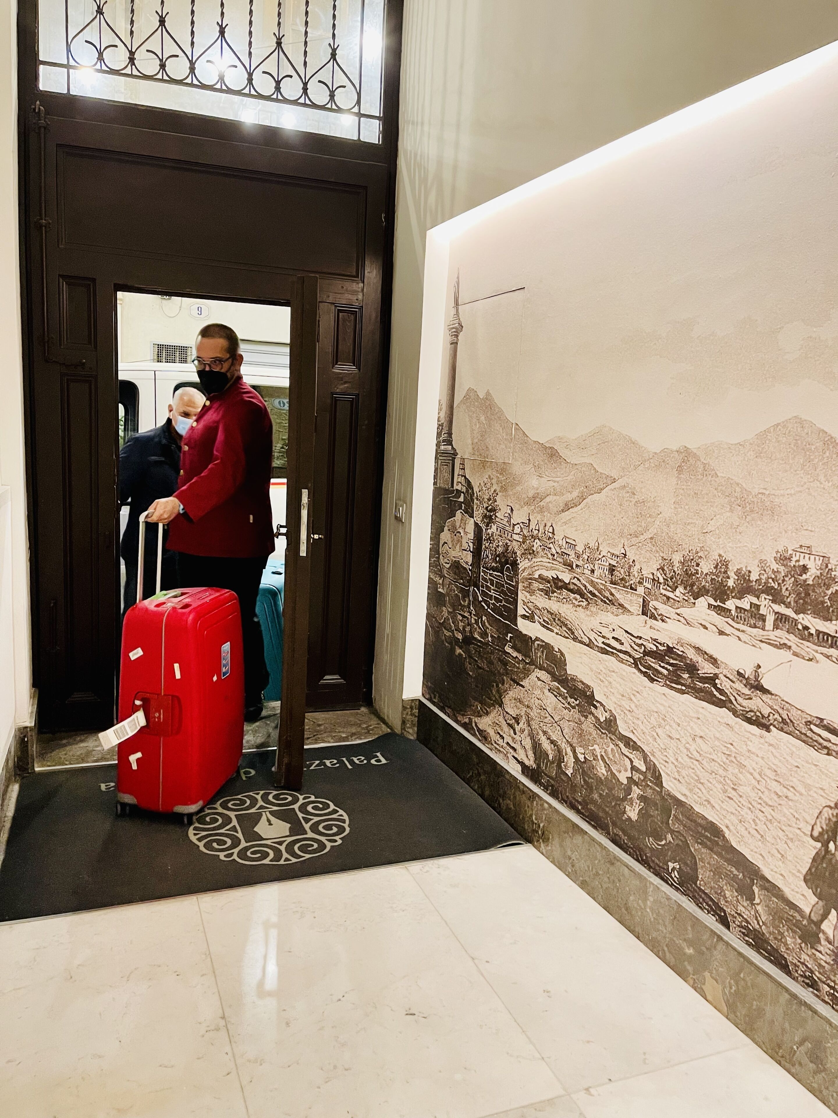 A hotel entrance with a large mural of a mountainous landscape, a red suitcase, and a person in a red jacket.