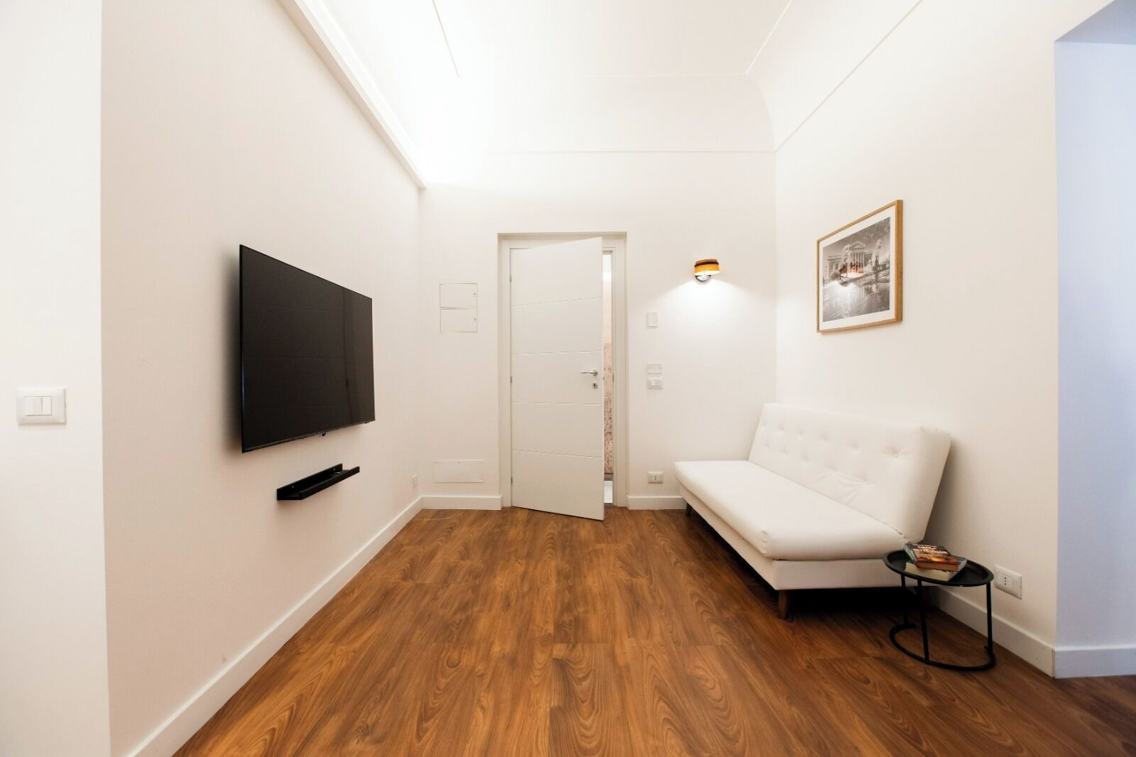 A hallway with a flat-screen TV mounted on the wall, a white sofa, and a framed picture on the wall.
