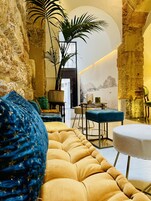 A cozy lounge area with a plush sofa, a small table, and chairs. The room features exposed stone walls and a large plant.