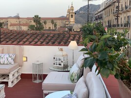 A rooftop terrace with a white sofa, a small table, and a lamp. In the background, there is a bell tower and buildings with red-tiled roofs.