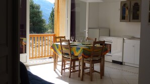 Un coin repas avec une table dressée pour quatre personnes, une cuisine avec des appareils électroménagers blancs et une vue sur les montagnes par une porte ouverte.