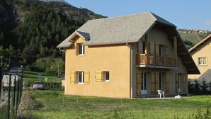 Une maison à deux étages dotée d’un balcon, entourée d’une pelouse et avec une montagne en arrière-plan.