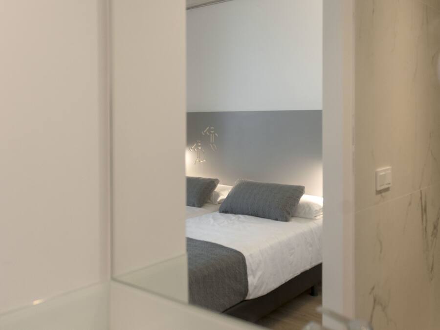 A modern bathroom with a large mirror, a white sink, and a visible bed in the reflection.