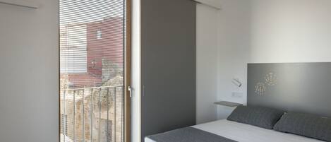 Un dormitorio con cama, ventana con persianas, luz en la pared y una pequeña estantería.