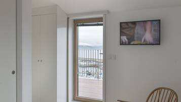 A room with a bed, a wooden chair, a television, and a view of the beach through a door.