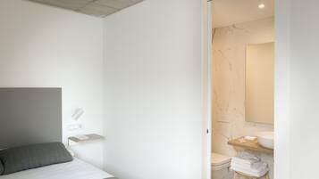 A modern bedroom with a bed, a bedside table, a wall-mounted light, and a bathroom with a toilet, sink, and towel rack.