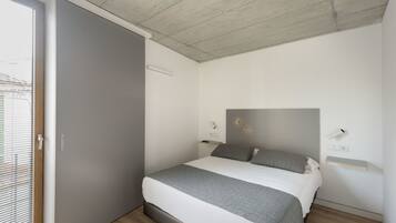 Un dormitorio con cama, ventana con persianas, techo con vigas a la vista y mesita de noche con lámpara.