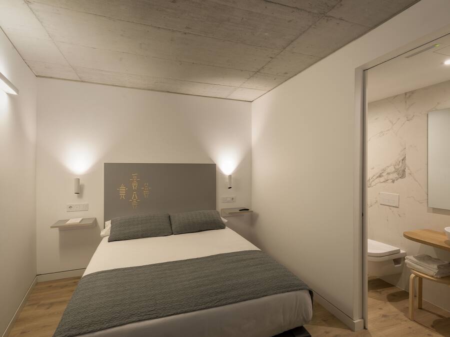 Un dormitorio moderno con cama, mesitas de noche, un espejo, un inodoro y un taburete de madera.