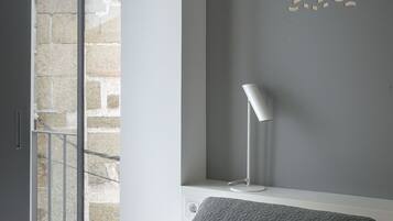 A bedroom with a bed, a bedside table, a lamp, and a view of a stone building through a window.