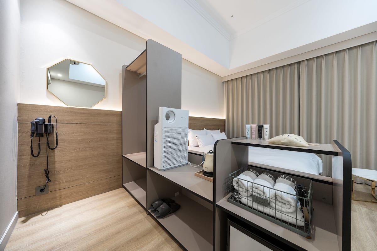A modern hotel room with a wooden accent wall, a large mirror, a desk with a computer, a bed with a white headboard, and a window with curtains.