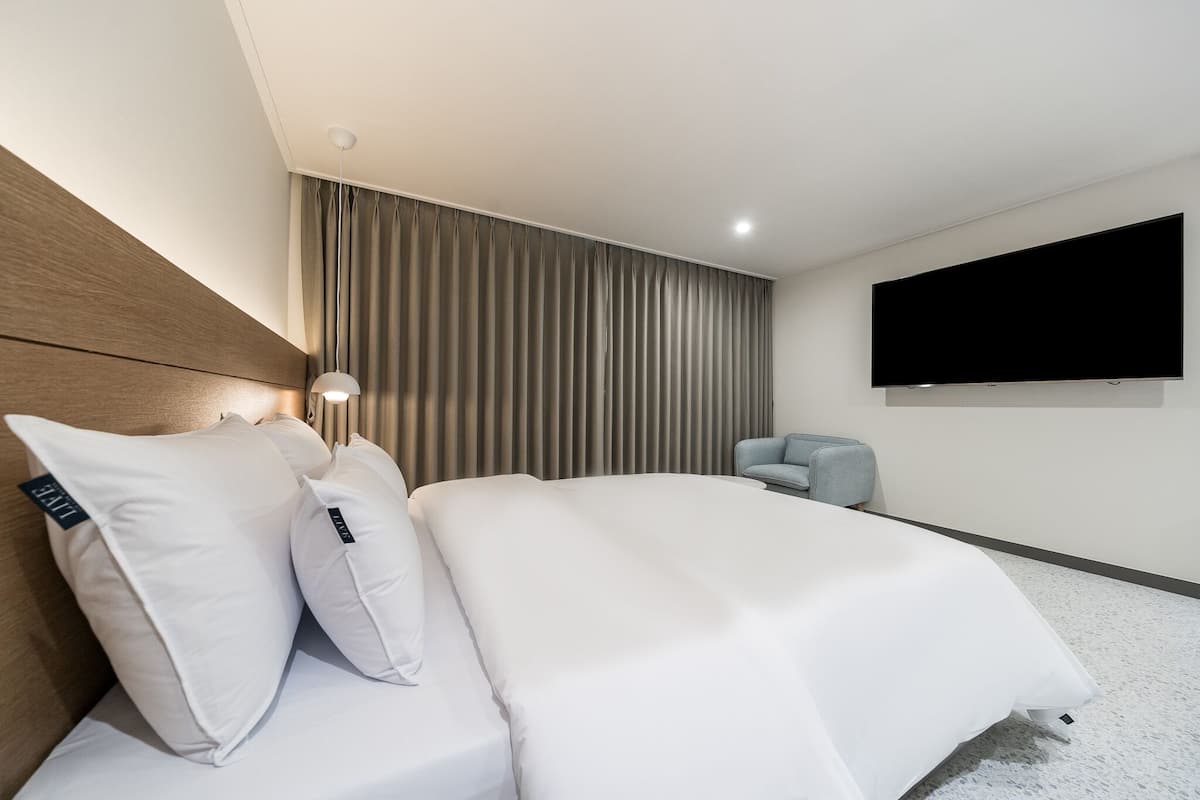 A modern hotel room with a large bed, a flat-screen TV, a wooden headboard, and a grey armchair.