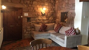 A cozy room with a stone wall, a wooden door, a grey sofa with striped pillows, a wooden table, and a patterned rug.