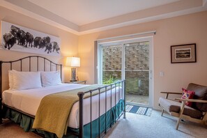 Ein Schlafzimmer mit Bett, Nachttisch, Lampe, einem Gemälde, einem Sessel und Blick durch eine Glastür.