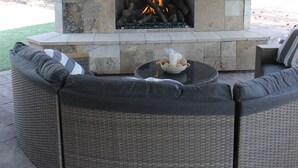 Ein Kamin aus Stein mit einem Fernseher darüber, ein Feuer brennt, und ein graues Wickersofa mit Kissen.