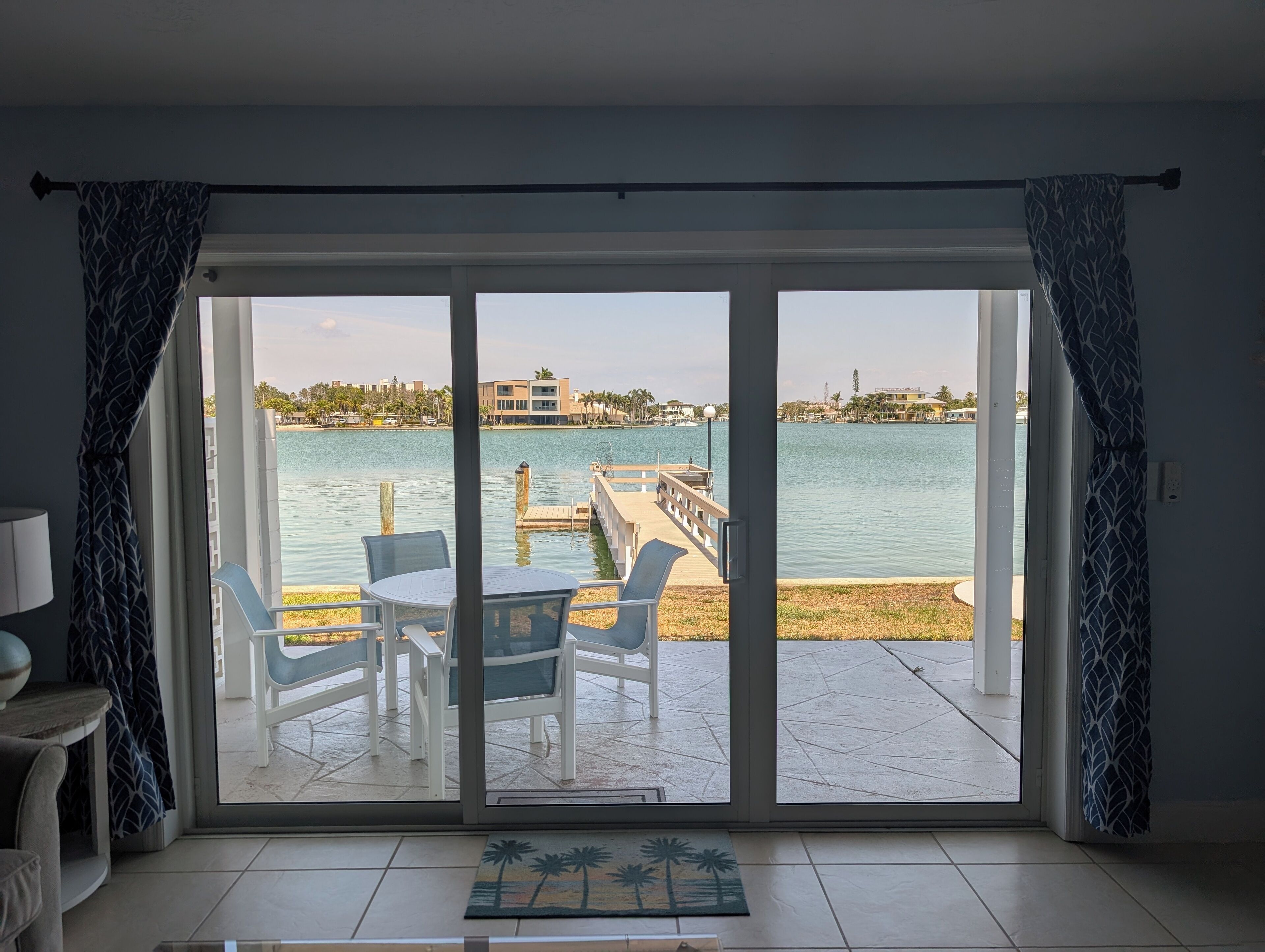A room with a sliding glass door leading to a patio with a table and chairs, a view of a body of water, and a distant building.