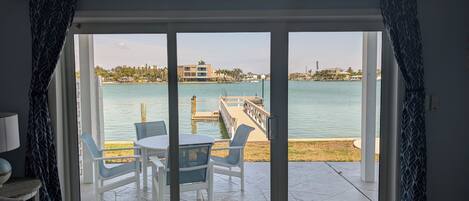 A room with a sliding glass door leading to a patio with a table and chairs, a view of a body of water, and a distant building.