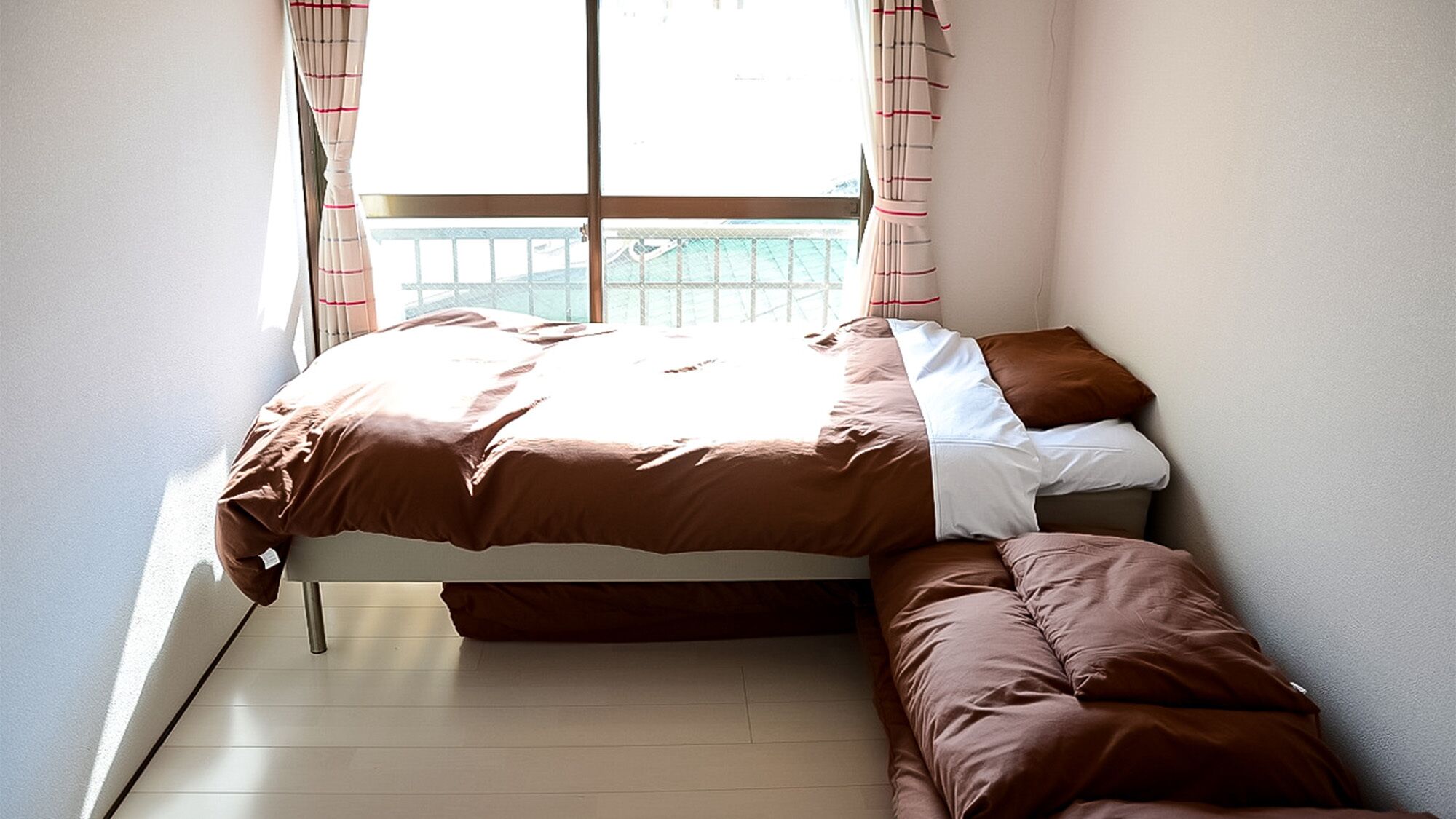 Een eenpersoonsbed met een bruine sprei en kussens, een bankje aan het voeteneind en een raam met gordijnen.