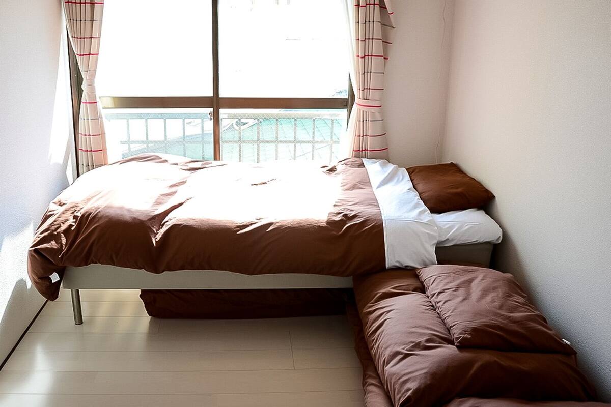 A single bed with a brown bedspread and pillows, a bench at the foot of the bed, and a window with curtains.