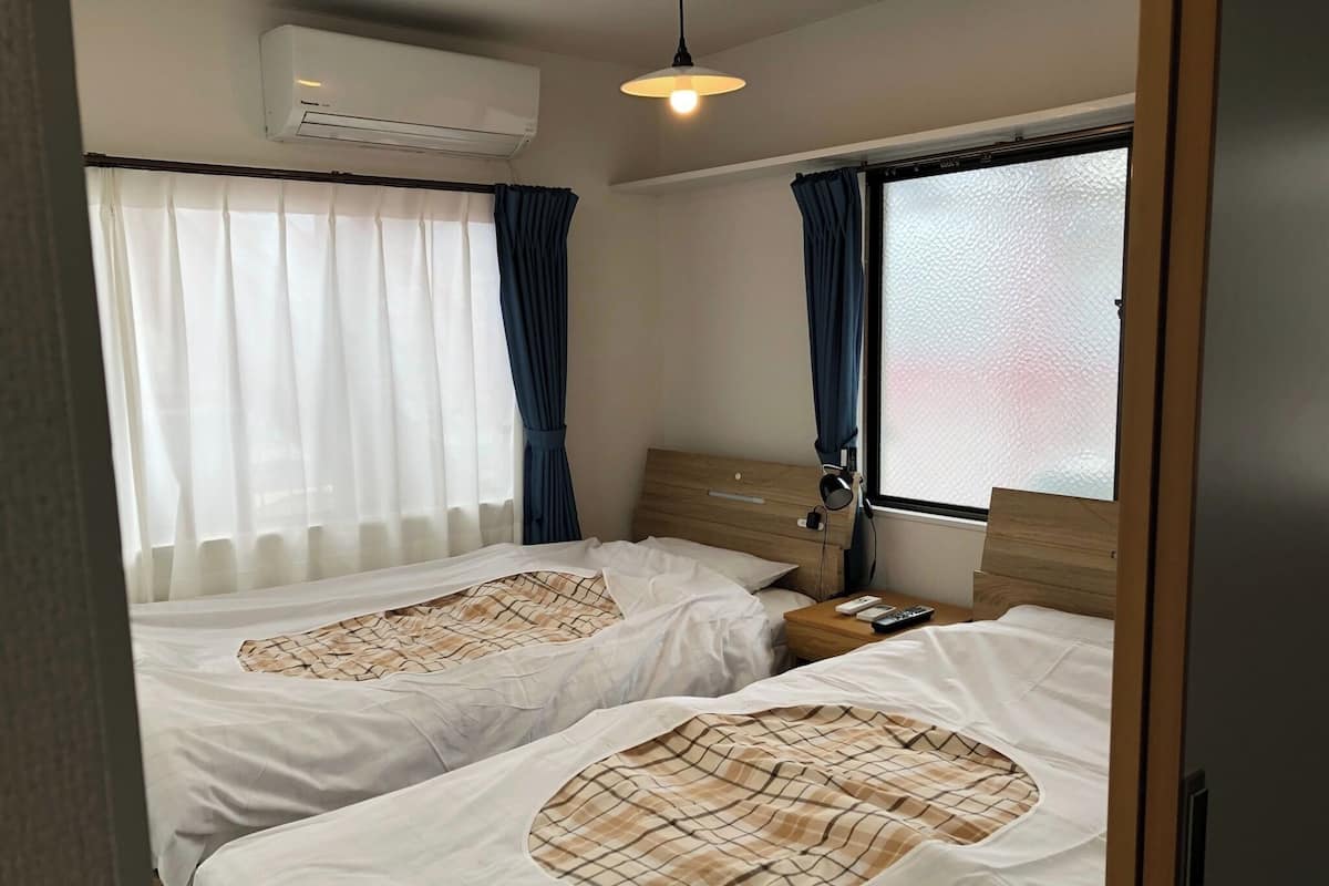 Habitación de hotel con dos camas, cabecero de madera, mesita de noche con lámpara y unidad de aire acondicionado en la pared.