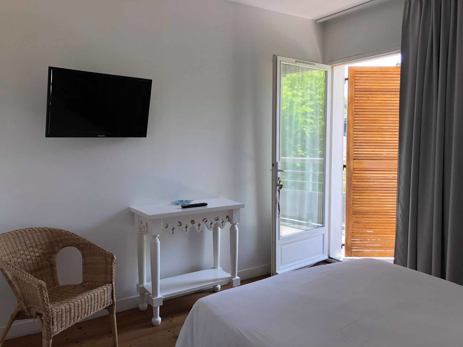 A bedroom with a bed, a television mounted on the wall, a small white table with a remote control, a wicker chair, and a door leading to a balcony with a view of greenery.
