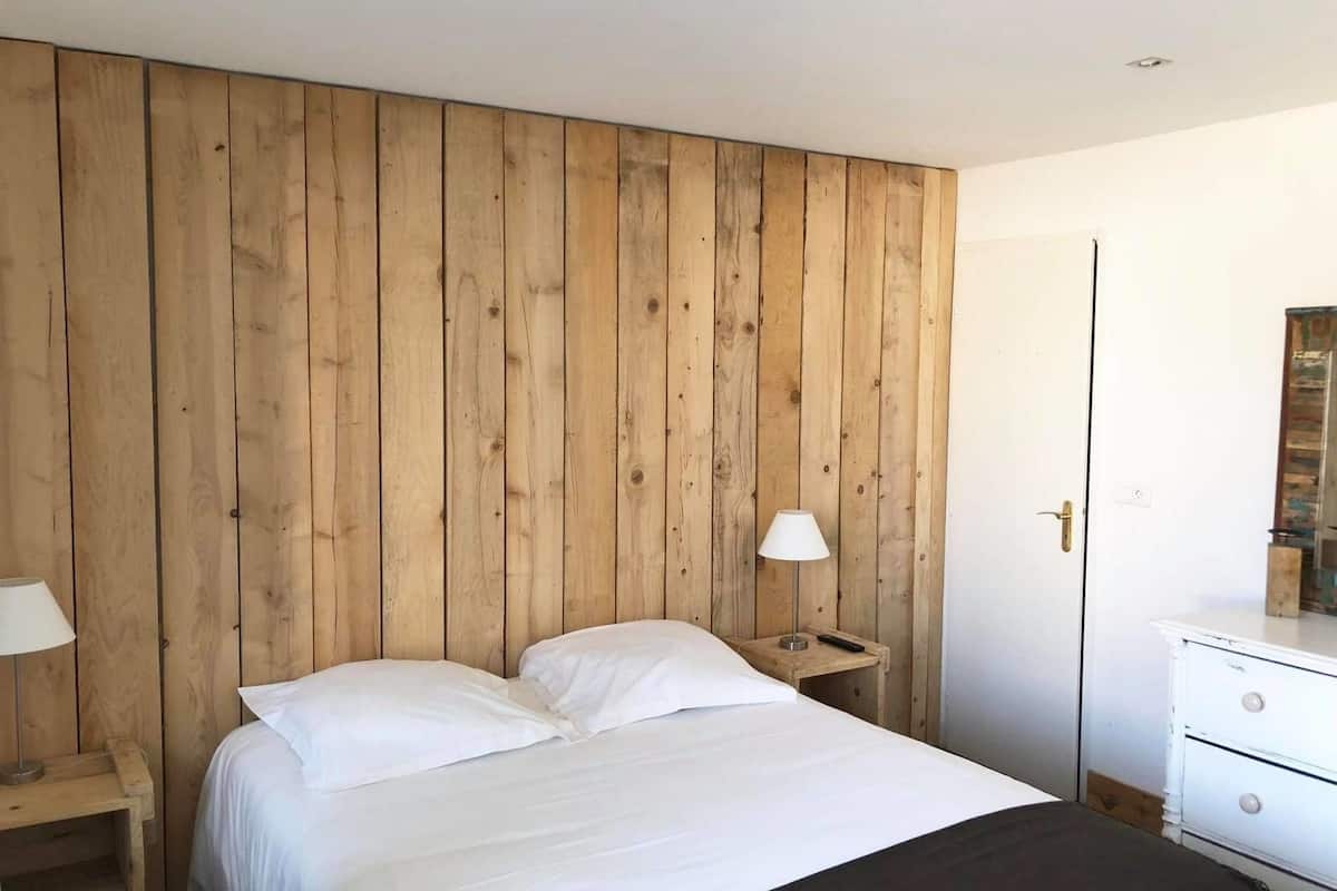 A bedroom with a wooden panel wall, a bed with white bedding, a bedside table with a lamp, and a dresser with drawers.