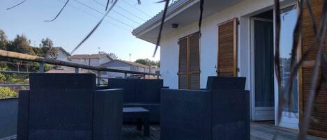 Uma área de estar coberta ao ar livre com móveis de vime escuro, um deck de madeira e um edifício com fachada branca e venezianas de madeira.