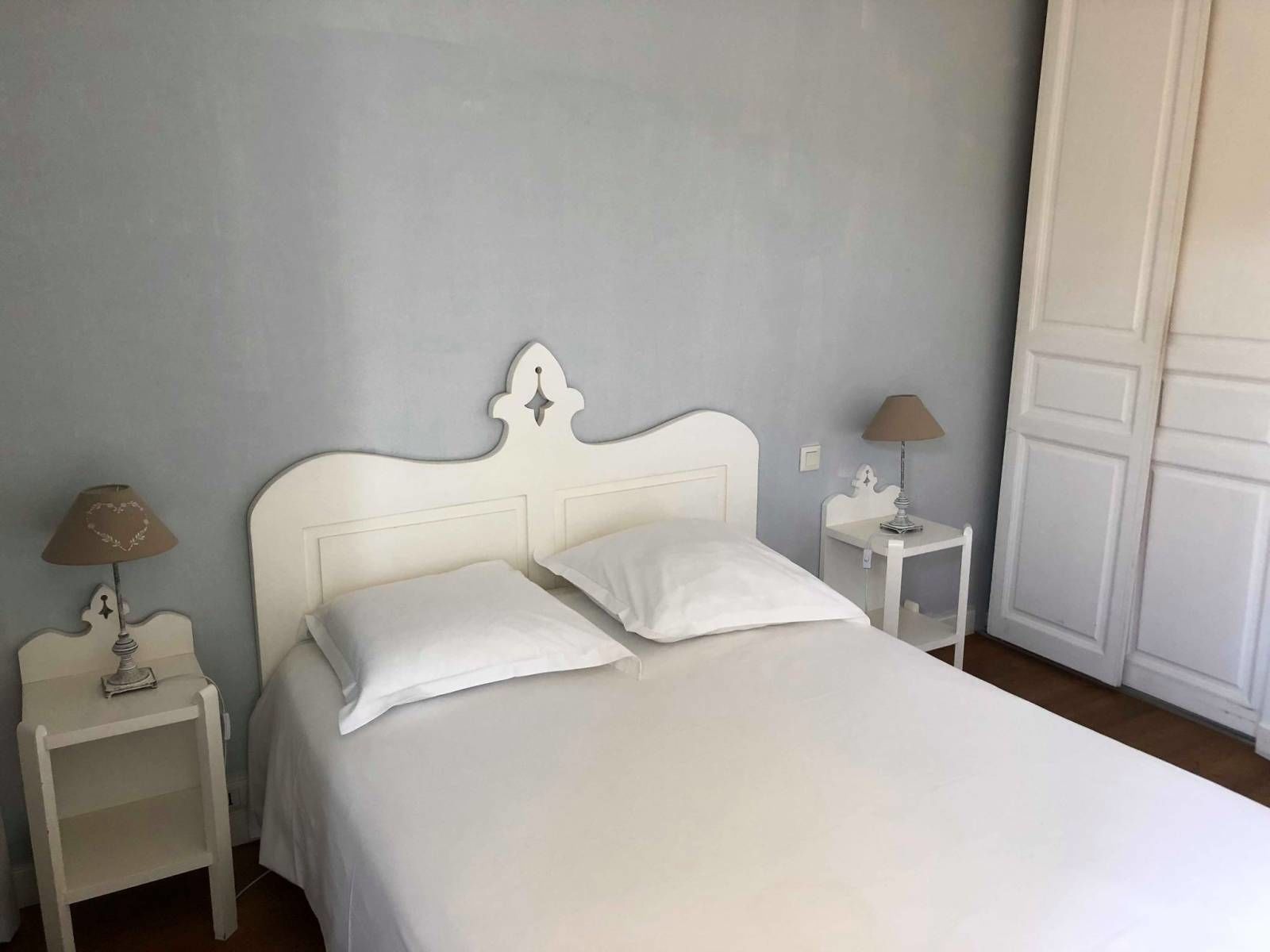 A neatly made bed with white bedding and pillows, two bedside tables with lamps, and a white wardrobe in a room with light-colored walls.
