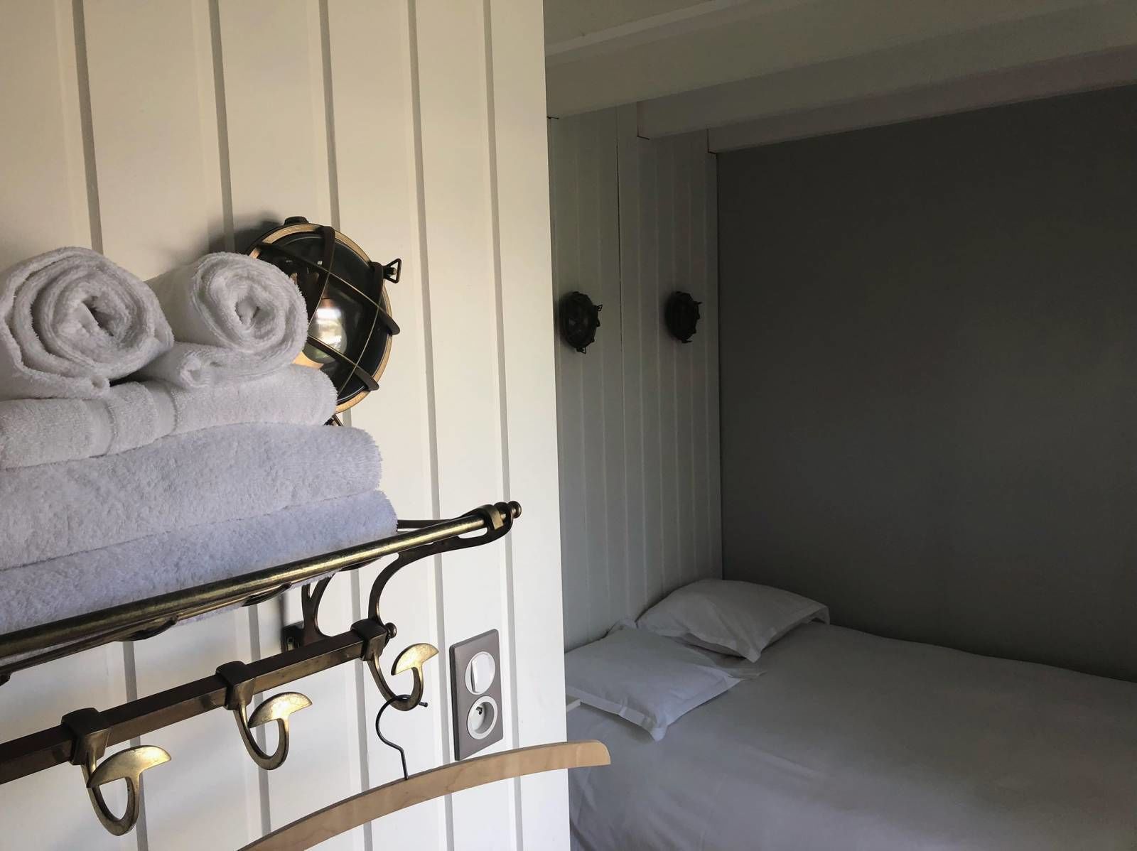 A neatly made bed with white linens, a wall-mounted towel rack with rolled towels, and a wall-mounted light fixture.