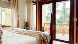 Een slaapkamer met een bed, gordijnen, een kamerplant en een balkon met een hekwerk.
