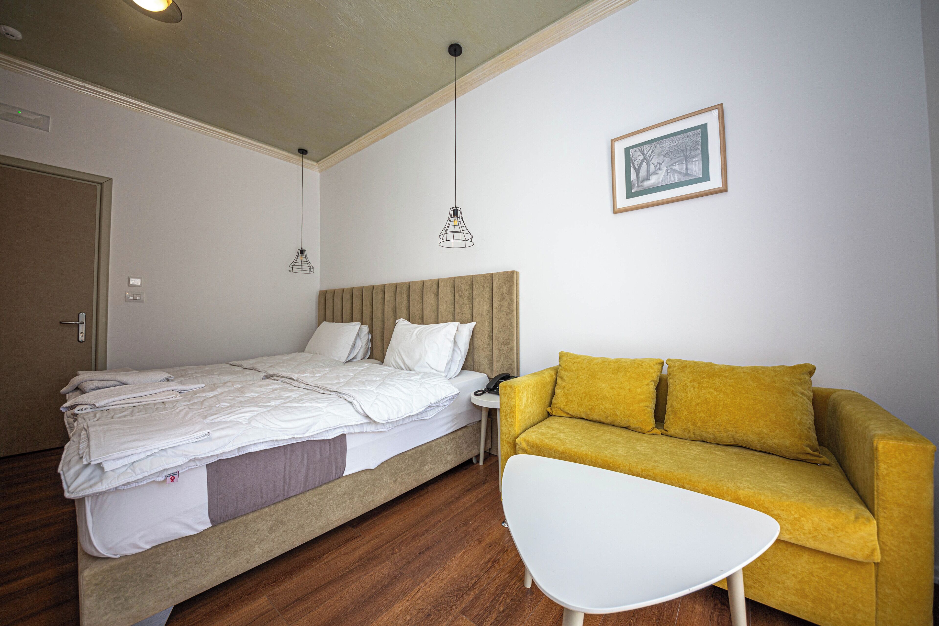 Een slaapkamer met een bed, een gele bank, een witte tafel en een lijst met een afbeelding aan de muur.