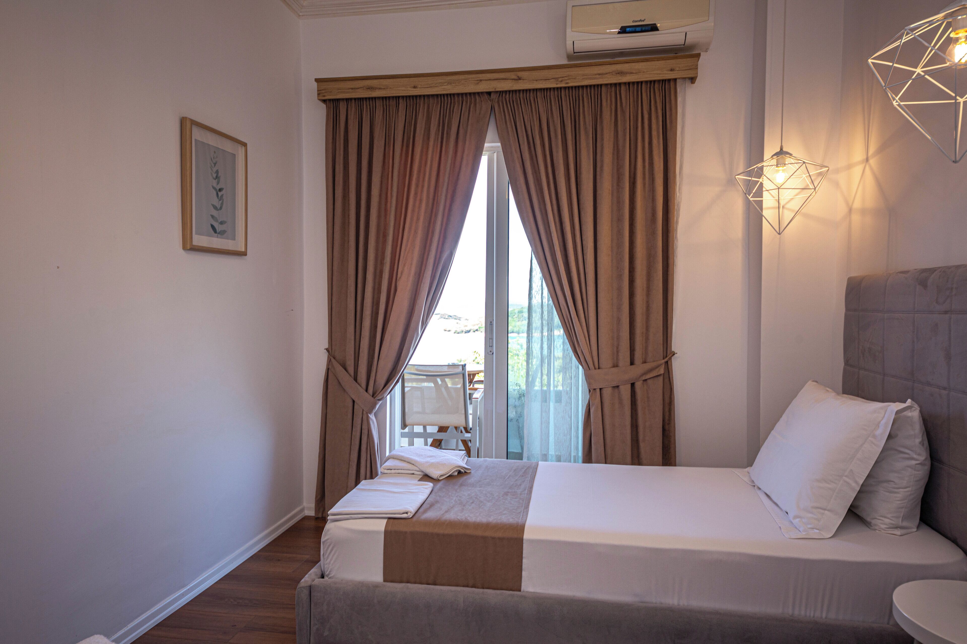 Una habitación de hotel con cama, ventana con cortinas, mesita de noche con lámpara y aire acondicionado instalado en la pared.