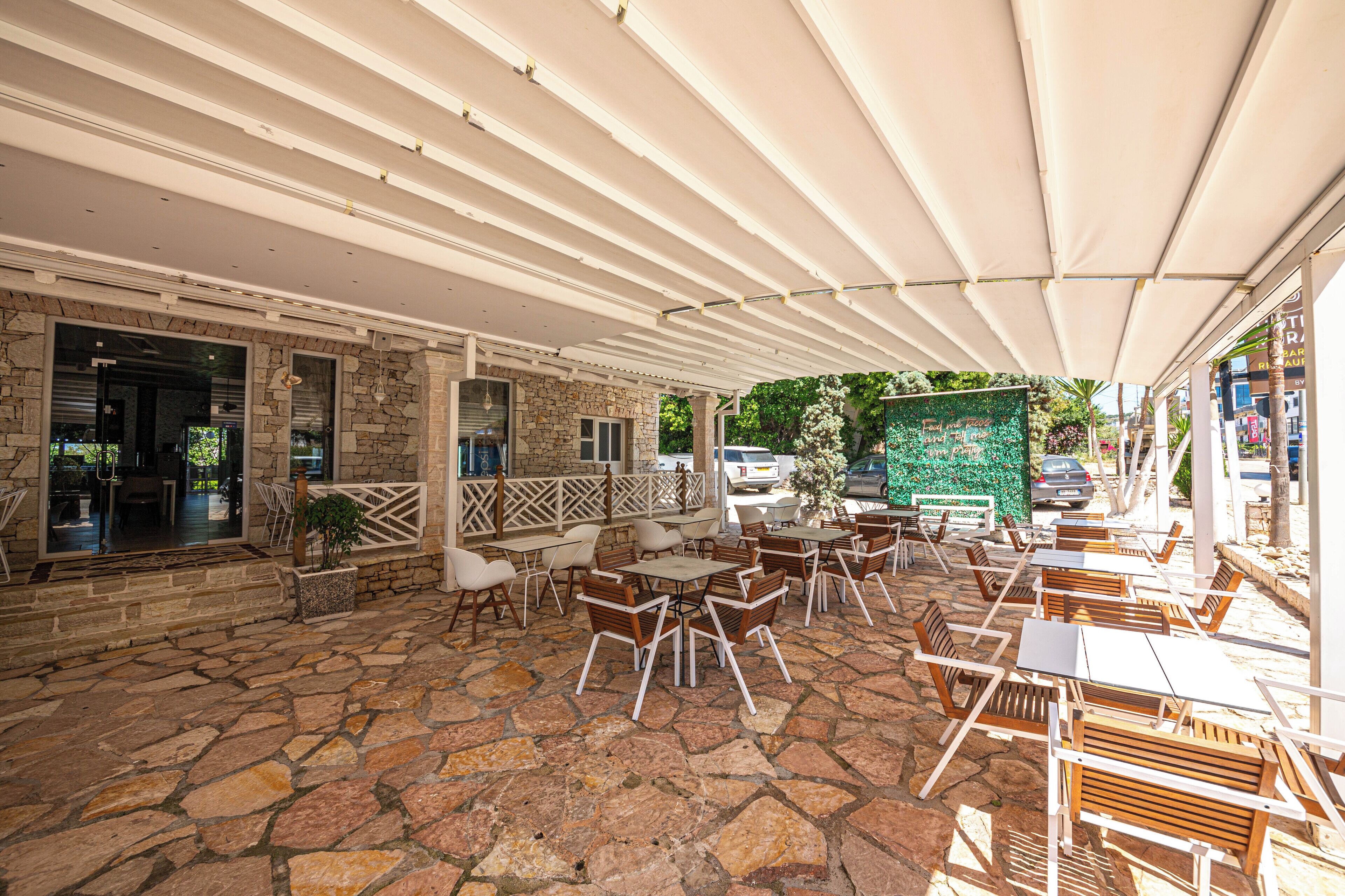 An outdoor cafe with stone flooring, wooden tables and chairs, and a covered area with a white canopy.