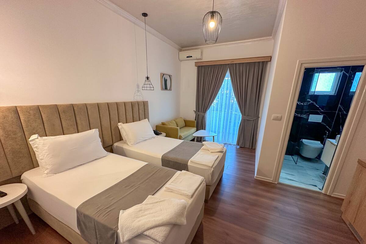 A hotel room with two beds, a small table, a chair, a sofa, and a view of the outside.