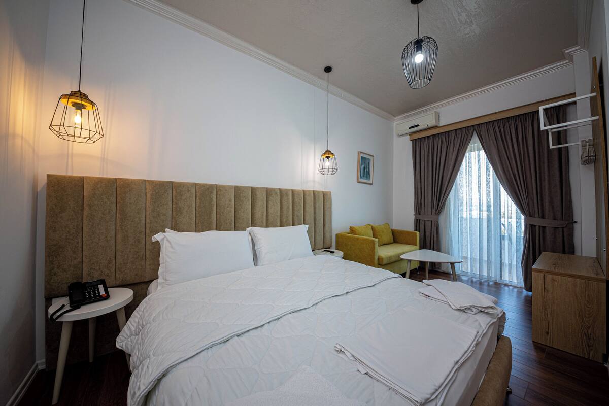 A hotel room with a large bed, a bedside table with a phone, a small table with a chair, a sofa, and a window with curtains.