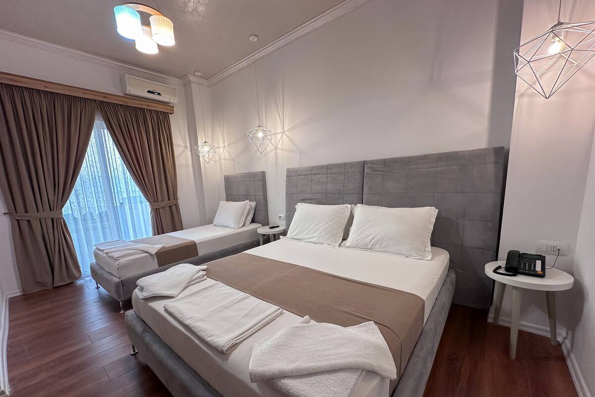 A modern hotel room with a large bed, two smaller beds, a bedside table, and a phone on a side table.