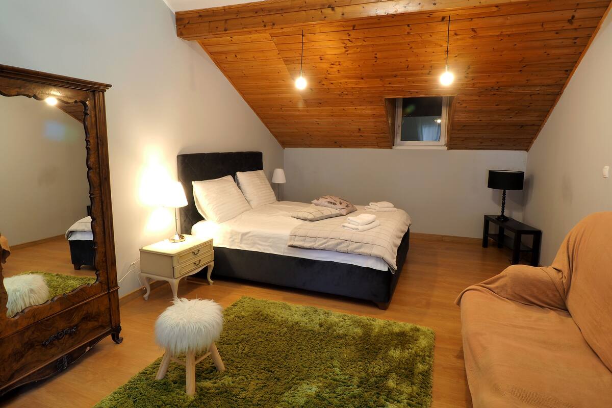 Ein Schlafzimmer mit schrägen Wänden, einem Bett mit weißer Bettwäsche, einem Nachttisch mit Lampe, einem Schminktisch und einem grünen Teppich. Ein Fenster ist ebenfalls zu sehen.