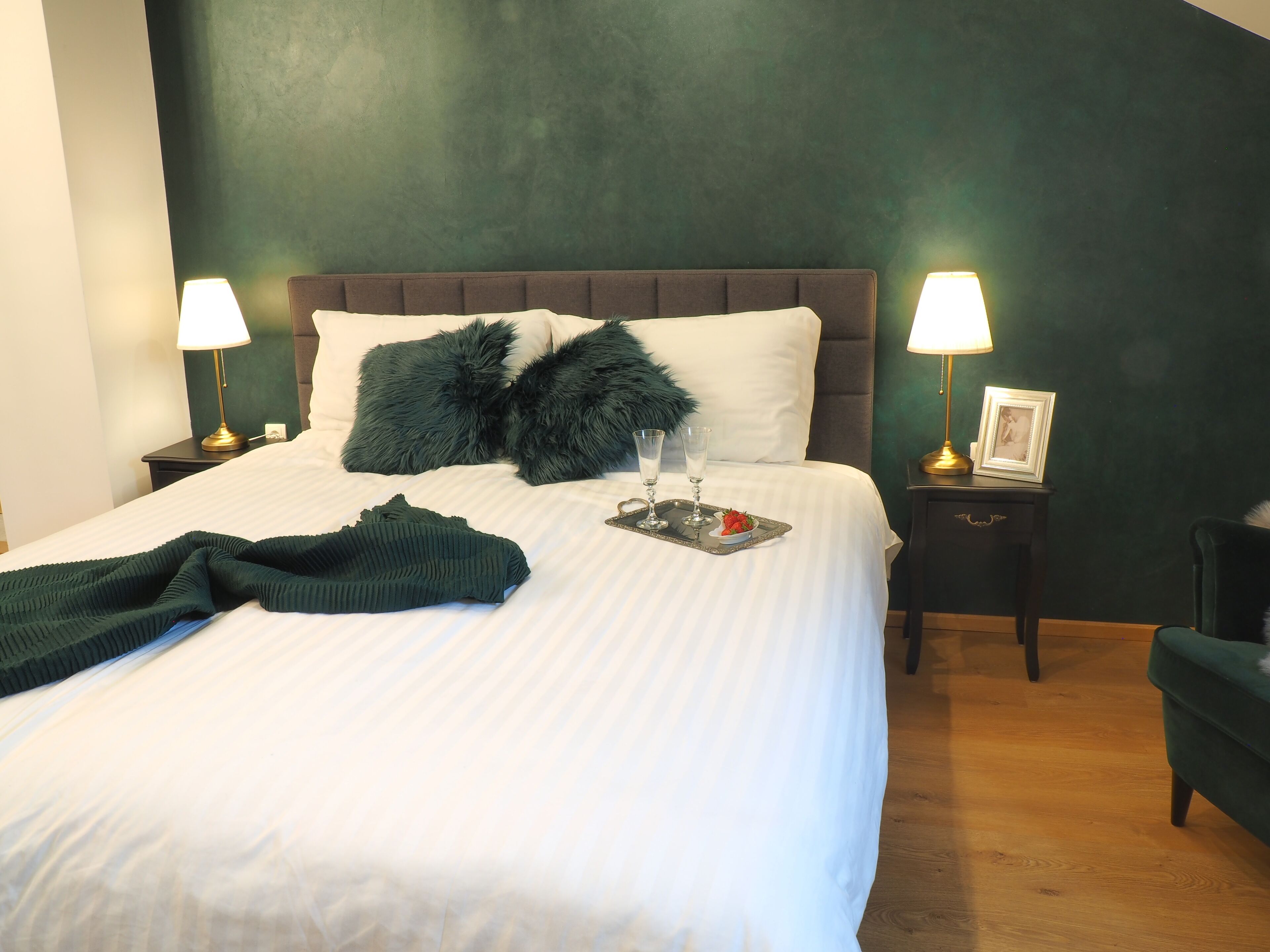 A neatly made bed with a dark green throw blanket, two fluffy pillows, and a tray with glasses and a strawberry. There are two bedside lamps and a small table with a picture frame.