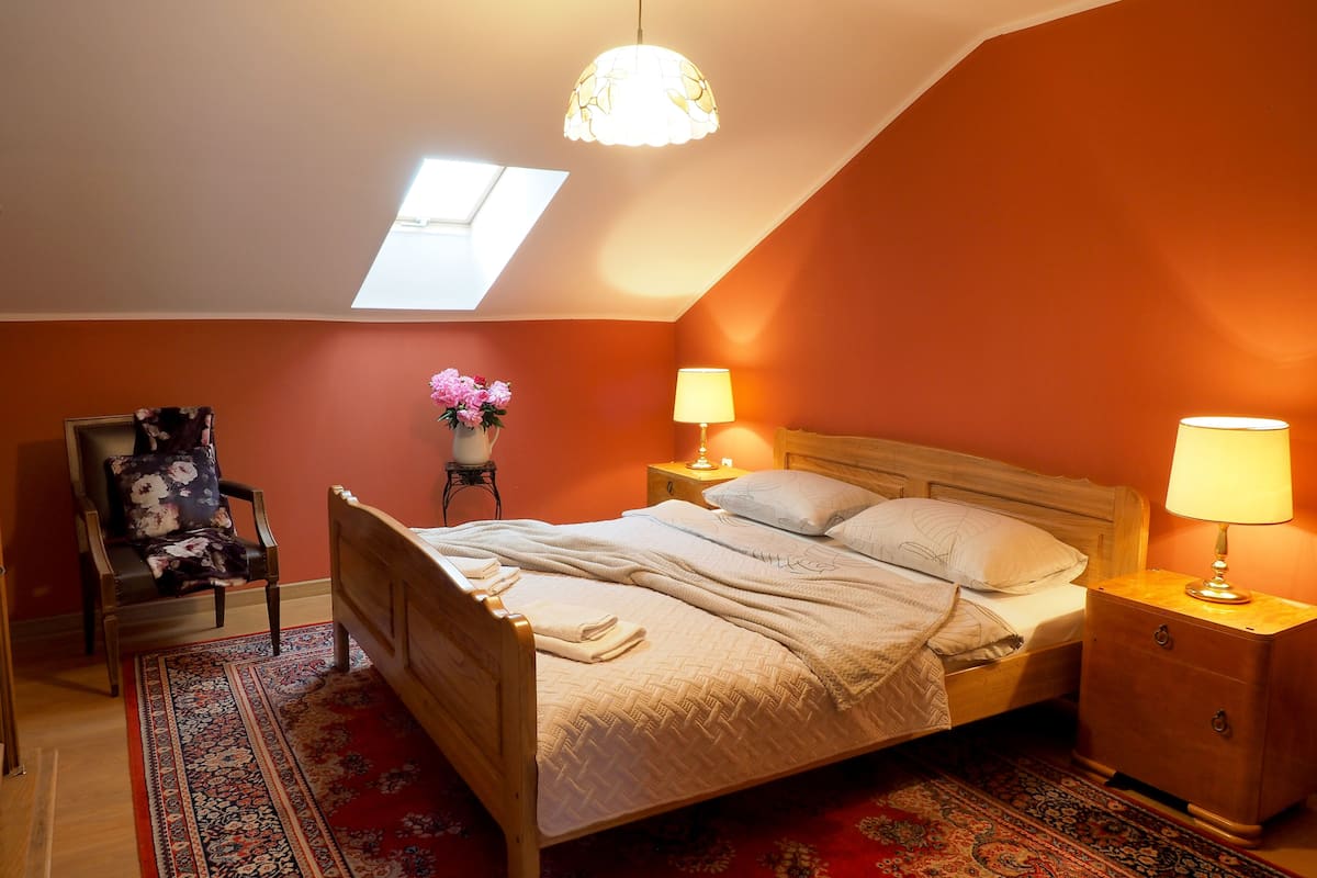 Ein Schlafzimmer mit schrägem Dach, einem Oberlicht, einem Bett mit Holzfußteil, zwei Nachttischen mit Lampen, einem Sessel mit Blumenkissen und einem gemusterten Teppich.