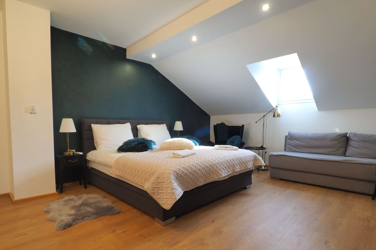 Ein modernes Schlafzimmer mit geneigter Decke, einem großen Bett mit dunklem Kopfteil, einem grauen Sofa und einem kleinen Tisch mit einer Lampe.