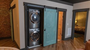A laundry room with a front-loading washer and dryer, a teal door, and a brick wall.