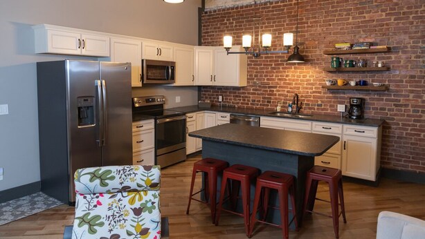A kitchen with a brick wall, white cabinets, a stainless steel refrigerator, a microwave, a coffee maker, and a bar area with stools.